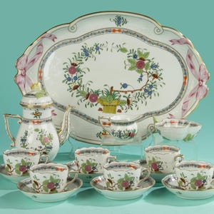 Könnte beinhalten: Ein komplettes Porzellan-Teeservice mit Blumen- und Bandmotiven. Das Set umfasst eine Teekanne, eine Servierplatte, Tassen, Untertassen, eine Zuckerdose und eine kleine Schüssel. Die Farbpalette umfasst weiße, goldene, rosa, blaue und grüne Akzente.
