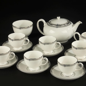 Op de afbeelding: Een wit theeservies bestaande uit een theepot, een melkkan en zes kopjes met bijpassende schotels. Elk onderdeel is versierd met een dunne zilveren rand en een subtiel grijs geometrisch patroon langs de rand. De set heeft een gladde afwerking.