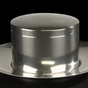 Op de afbeelding: Een gepolijste zilverkleurige cilindrische houder met een plat deksel, rustend op een brede, ronde basisplaat. De basisplaat is voorzien van een delicaat detail met een kralenrand. Het gehele object heeft een gladde, reflecterende afwerking.