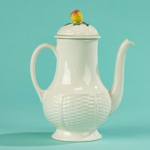 Può includere: Caffettiera in ceramica bianca con un coperchio che presenta un piccolo disegno di frutta gialla. La caffettiera ha un motivo a cesto intrecciato e un beccuccio laterale.