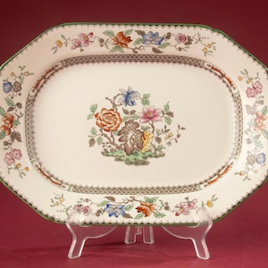Puede incluir: Una bandeja de servicio ovalada de cerámica con base color crema, que presenta un motivo floral central en naranja, rosa, azul y verde. El borde está decorado con un patrón de enredaderas florales, un filo verde oscuro y una cenefa de rejilla marrón.
