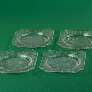 Op de afbeelding: Vier vierkante platen van helder glas met een decoratief geëtst ontwerp. De platen zijn in een vierkant patroon op een groene achtergrond gerangschikt.