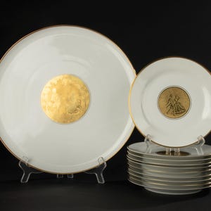 Puede incluir: Un juego de platos de porcelana blanca con bordes dorados y un diseño de medallón dorado en el centro. El juego incluye platos grandes y platos más pequeños apilados en un soporte transparente, sobre un fondo negro.