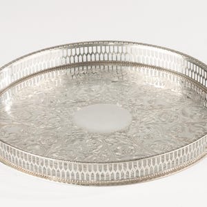 Könnte beinhalten: Ein rundes silberfarbenes Serviertablett mit einem komplizierten geprägten Blumenmuster auf der Basis und einem hohen, durchbrochenen Galerierand mit einem sich wiederholenden vertikalen Gitterdesign. Die Mitte des Tabletts bleibt glatt.