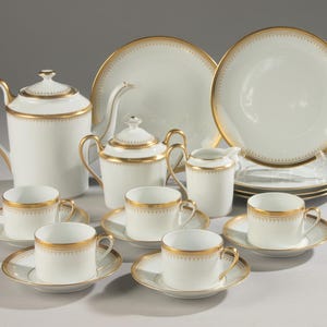 May include: A white porcelain tea set featuring a gold rim with a delicate repeating pattern. The set includes a teapot, a lidded sugar bowl, a creamer, five teacups with matching saucers, and two stacked plates, all with a smooth, glossy finish.