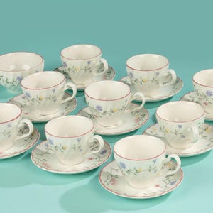 Puede incluir: Un juego de tazas y platillos blancos con un diseño floral y un borde rojo. El juego incluye un cuenco. Las tazas y platillos están dispuestos sobre una superficie azul claro. Las tazas tienen aproximadamente 7.6 cm de diámetro.