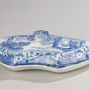 Può includere: Un piatto da portata in porcellana blu e bianca con coperchio. Il piatto ha una forma unica ed è decorato con una scena paesaggistica dettagliata. Il coperchio è dotato di manico e il bordo presenta un motivo floreale.