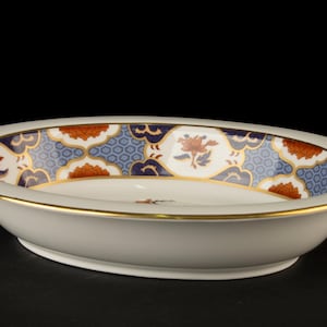 Puede incluir: Cuenco de cerámica blanco de forma ovalada con borde decorativo. El borde presenta un patrón floral azul, naranja y blanco con detalles dorados. El cuenco tiene un ribete dorado.