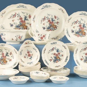 May include: A large dinnerware set featuring cream-coloured ceramic pieces with scalloped edges and a floral pattern in shades of blue, pink, and brown. The collection includes various plates, bowls, a gravy boat, and a serving platter with floral motifs.