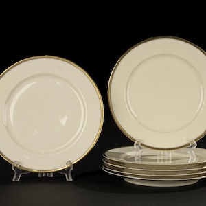 Può includere: Un set di piatti da cena color crema con bordo dorato e bordo dorato a pois. I piatti sono esposti su supporti di plastica trasparente su sfondo nero. Il set comprende cinque piatti.