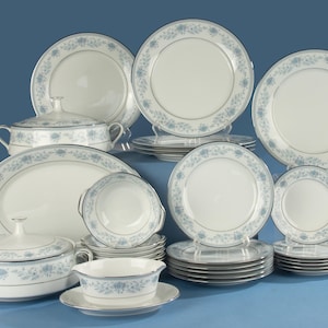 Op de afbeelding: Een servies set van wit porselein met een delicaat blauw bloemenpatroon langs de randen en een zilveren afwerking. De collectie bevat diverse borden, kommen, serveerschalen met deksel en een sauskom met bijpassende schotel.
