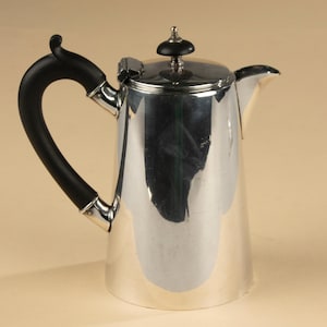 Puede incluir: Una cafetera plateada pulida con un asa y un pomo de tapa negros. La cafetera tiene un asa curvada y un pico para verter. La tapa es abatible y tiene un pequeño pomo decorativo. La altura es de unos 20 cm.