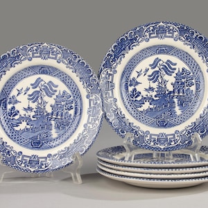 Op de afbeelding: Een set blauw-witte keramische borden met een Willow-patroon. De borden tonen een centrale landschapsafbeelding met een pagode, brug en vogels. De set bevat twee borden op standaards en een stapel van vier borden.