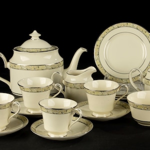 Op de afbeelding: Een crèmekleurige theeset met een decoratief bloemmotief en zilveren rand. De set bevat een theepot, melkkan, suikerpot, kopjes, schotels en een bord. De theeset wordt gepresenteerd op een zwart oppervlak.