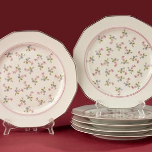 Op de afbeelding: Een set witte, achthoekige borden met een delicaat bloemmotief in roze en blauw. Elk bord heeft een roze rand en een zilveren rand. De set bevat borden op standaards en een stapel borden.