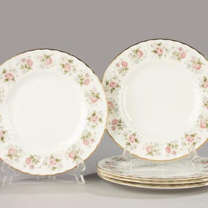 May include: A set of white porcelain plates with a floral rose pattern in pink and green. The plates have a gold trim and a scalloped edge. Several plates are stacked on top of each other.