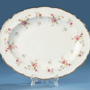 May include: An oval porcelain serving platter featuring a white base with a delicate floral pattern of pink roses, yellow buds, and small blue flowers. The platter has a scalloped edge finished with a thin, metallic gold trim along the rim.