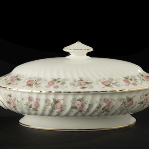 May include: An oval porcelain tureen with a lid, featuring a cream-coloured base with a ribbed texture. The exterior is decorated with a repeating pattern of small pink roses and green leaves, accented by a thin gold trim along the edges and handles.