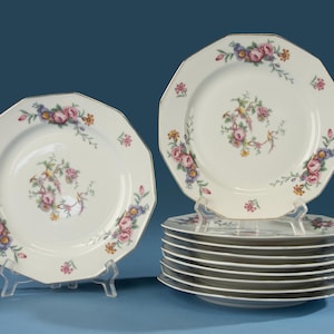 Op de afbeelding: Een set achthoekige witte keramische borden met een centraal bloemmotief van roze rozen, blauwe bloemen en kleine gele bloesems. De rand is versierd met bijpassende bloemmotieven en een dunne metalen rand, gepresenteerd in een stapel.