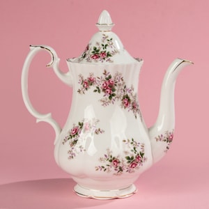 Op de afbeelding: Een witte porseleinen theepot met een bloemenpatroon van roze rozen en groene bladeren. De theepot heeft een gouden rand rond de deksel en de basis.