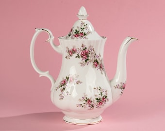 Pode incluir: Uma chaleira de porcelana branca com um padrão floral de rosas cor-de-rosa e folhas verdes. A chaleira tem uma borda dourada em torno da tampa e da base.