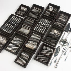 May include: A collection of silver-colored flatware sets, including forks, knives, spoons, and serving utensils. The silverware is arranged in black boxes, with some pieces displayed separately. The utensils have a reflective, polished finish.