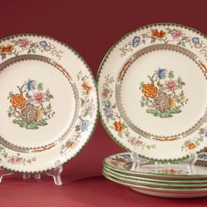 Può includere: Un set di sei piatti rotondi in ceramica con base color crema e un motivo floreale centrale in arancione, rosa e blu. I piatti presentano un bordo geometrico marrone e un bordo smerlato verde, con ulteriori motivi floreali sul bordo.