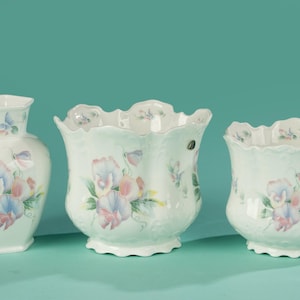 May include: Three white ceramic flower pots with a floral design. The pots are different sizes and shapes. The design features pink, blue, and white flowers with green leaves.