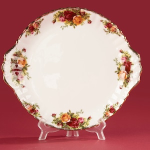 Op de afbeelding: Een witte serveerschaal met geschulpte rand en een bloemenpatroon van rozen in tinten rood, geel en roze. De schaal heeft een gouden rand en wordt gepresenteerd op een transparante plastic standaard. De schaal is waarschijnlijk gemaakt van porselein of keramiek.