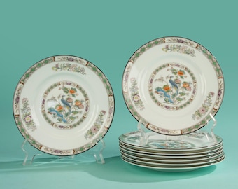 May include: A set of white ceramic plates with a decorative peacock and floral design. The plates have a black rim and a green and floral border. The set includes plates on clear stands and a stack of plates.