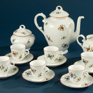 Könnte beinhalten: Ein weißes Porzellan-Teeservice mit floralen Mustern. Das Set beinhaltet eine Teekanne, eine Zuckerdose, einen Sahnekännchen und sechs Tassen mit Untertassen. Jedes Teil hat einen roten Rand und ist mit bunten Blumen verziert.