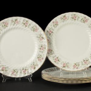 May include: A set of six white ceramic plates featuring a scalloped edge and a delicate floral pattern of pink roses and small blue flowers. Each plate has a gold-trimmed rim and a subtle spiral texture radiating from the centre to the edge.