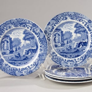 Può includere: Un set di piatti decorativi blu e bianchi. Ogni piatto presenta una scena paesaggistica con edifici, alberi e figure. I piatti hanno un bordo floreale dettagliato. Il set include piatti su supporti trasparenti.