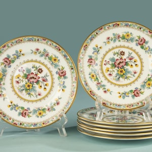 Pode incluir: Um conjunto de seis pratos de cerâmica branca com um bouquet floral central em tons de rosa, amarelo e azul, rodeado por uma borda floral correspondente e um acabamento dourado. Os pratos estão expostos em suportes transparentes.
