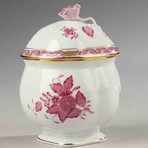 Op de afbeelding: Een witte porseleinen suikerpot met deksel, versierd met roze bloemmotieven en gouden randen. Het deksel heeft een roze roos en bladeren. De kom heeft een afgeronde body en een vierkante basis.