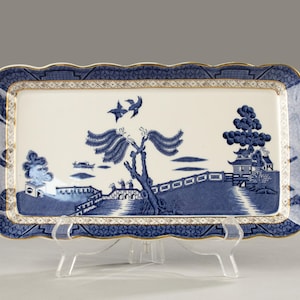 Op de afbeelding: Een rechthoekige blauw-witte porseleinen serveerschaal met een gouden rand. De schaal heeft een Willow-patroonontwerp met een brug, bomen en vogels. De randen hebben een geschulpte vormgeving.