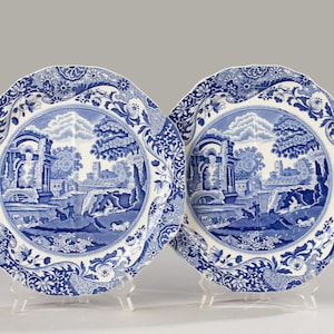 Puede incluir: Dos platos decorativos azules y blancos con un paisaje. Los platos tienen un borde festoneado y un borde floral detallado. La imagen central representa un paisaje clásico con edificios, árboles y figuras.