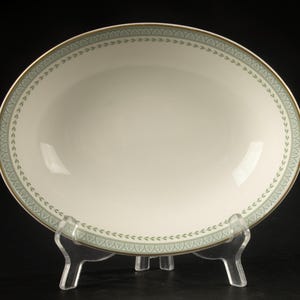 May include: An oval-shaped ceramic bowl with a cream-colored center and a wide border featuring a sage green pattern of repeating leaf-like motifs. The rim is finished with a thin gold-colored band, and the bowl is displayed on a clear stand.