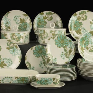 Op de afbeelding: Een grote serviesset met een crèmekleurige basis en een herhalend bloemmotief in tinten blauwgroen en olijfgroen. De collectie bevat diverse ronde borden, kommen, serveerschalen met deksel en een ovale serveerschaal met handgrepen.