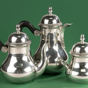 May include: A set of three silver-plated serving pieces, including a teapot, a coffee pot, and a creamer. The teapot and coffee pot have black handles. The pieces are displayed against a green background.