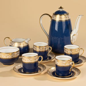 Op de afbeelding: Een marineblauwe koffieset met gouden details, waaronder een koffiepot, kopjes, schotels, een melkkan en een suikerpot. De set heeft decoratieve gouden patronen. Gepresenteerd op een lichtbeige oppervlak.