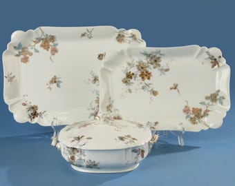 May include: A white porcelain serving set featuring floral designs in muted tones of brown, pink, and blue. The set includes two rectangular platters with scalloped edges and a covered serving dish. The pieces are displayed on clear stands against a blue backdrop.