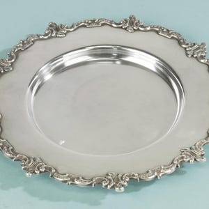 Op de afbeelding: Een zilverkleurige plaat met een sierlijke, decoratieve rand. De plaat heeft een brede rand met een gedetailleerd, scrollend ontwerp. Het midden van de plaat is glad en reflecterend, geschikt voor het serveren van voedsel.