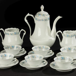 May include: A white porcelain tea set with a floral design. The set includes a teapot, creamer, sugar bowl, and several teacups with saucers. The floral pattern features blue and light green accents. The set is arranged on a dark surface.
