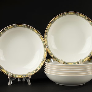 May include: A set of white ceramic bowls with a decorative border featuring gold, blue, and green geometric patterns. The bowls are displayed on clear plastic stands against a black background. The set includes several bowls.