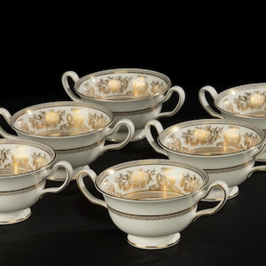 Puede incluir: Seis tazas de sopa de porcelana blanca con diseños florales dorados y ribetes dorados. Cada taza tiene dos asas y una base de pedestal. Las tazas están dispuestas sobre una superficie negra.