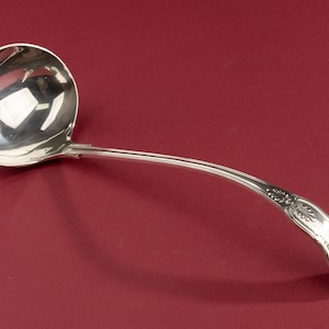 May include: A polished silver-toned metal soup ladle featuring a deep, rounded bowl and a long, curved handle. The handle is decorated with an embossed shell-like motif at the tip and along the stem, showcasing a reflective, metallic finish.
