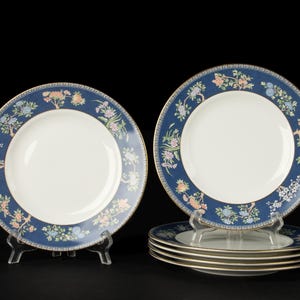 Puede incluir: Un juego de platos redondos de cerámica con centro blanco y un borde ancho azul oscuro con patrón texturizado. El borde está decorado con ramas florales coloridas y un borde dorado, complementado por un diseño geométrico de grecas.