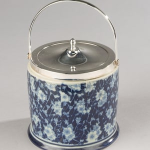 Op de afbeelding: Een cilindrische keramische ijsemmer met een donkerblauw en wit bloemmotief over de buitenkant. De houder is afgewerkt met een gepolijst zilverkleurig metalen deksel en een bijpassend gebogen handvat, inclusief een kleine centrale knop.