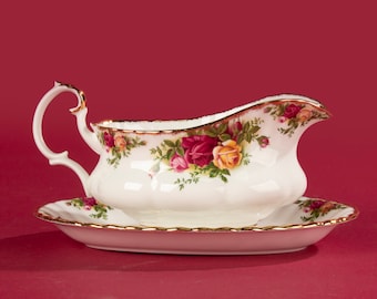 Può includere: Una salsiera in porcellana bianca con bordo dorato e un motivo floreale con rose rosse, gialle e rosa. La salsiera è su un sottopiatto abbinato.